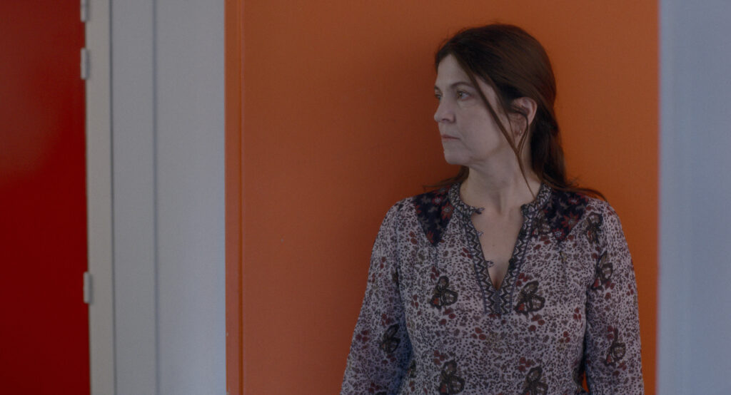 ine Frau mit dunklem, zurückgebundenem Haar steht in einem Innenraum vor einer orangefarbenen Wand und blickt nach rechts. Sie trägt eine gemusterte Bluse und wirkt nachdenklich.