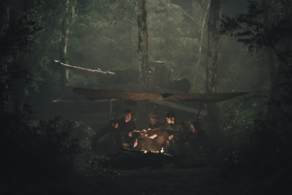 Eine Gruppe von Soldaten sitzt nachts in einem düsteren Wald im Schein eines Lagerfeuers zusammen. Im dunklen Hintergrund ist die Silhouette eines Panzers (Tiger) unter einer Plane zwischen Bäumen zu erkennen.