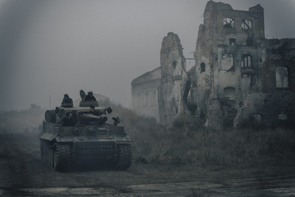 Szene aus Der Tiger: Ein historischer Panzer (Tiger) fährt an einer zerstörten, verlassenen Gebäuderuine vorbei. Das Szenario ist in dichtem Nebel und trüben, grauen Farbtönen gehalten, während Soldaten aus den Luken des Panzers schauen.