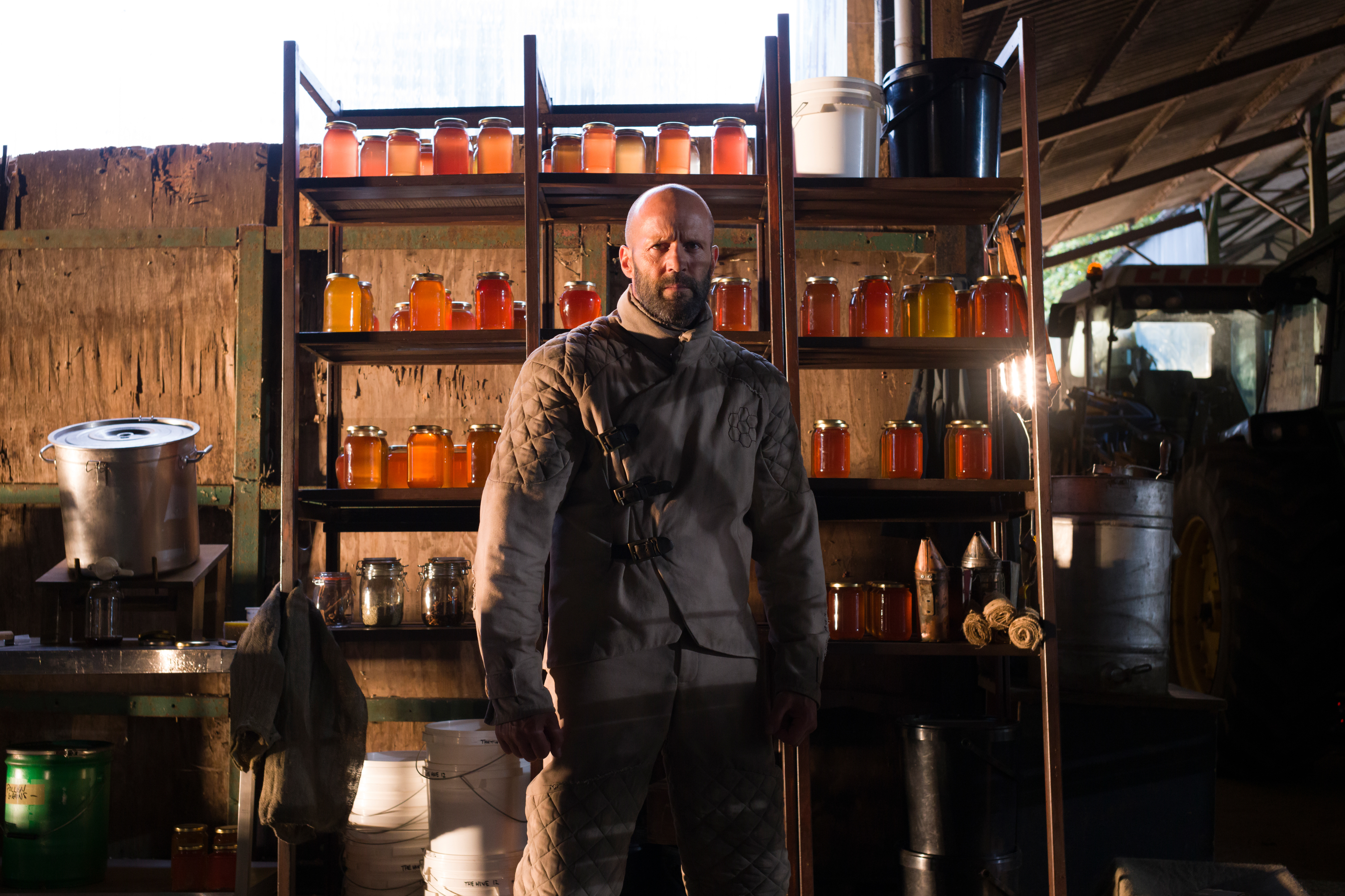 Filmszene aus dem Actionfilm „The Beekeeper“ mit dem Hauptdarsteller Jason Statham als Adam Clay. Er steht als glatzköpfiger, bärtiger Mann im Zentrum des Bildes in einer Ganzkörperaufnahme vor einem großen Holzregal in einer ländlichen Scheune. Er trägt einen hellgrauen, gesteppten Imker-Schutzanzug und blickt mit ernstem, entschlossenem Gesichtsausdruck direkt nach vorne. Das Regal hinter ihm ist dicht bestückt mit zahlreichen Einmachgläsern, die mit goldgelbem und bernsteinfarbenem Honig gefüllt sind. Im rustikalen Hintergrund sind Eimer, ein großer Kochtopf sowie die Silhouette eines Traktors zu erkennen.