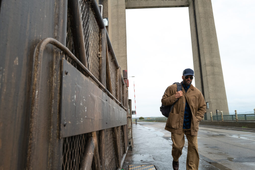 Filmszene aus dem Actionfilm „The Beekeeper“. Der Hauptdarsteller Jason Statham läuft im rechten Bildvordergrund eine nasse, asphaltierte Straße entlang, die über eine Brücke führt. Er trägt eine braune Arbeitsjacke, passende braune Hosen, eine dunkelblaue Basecap und eine Sonnenbrille, während er eine dunkle Umhängetasche über der Schulter trägt. Im linken Vordergrund verdeckt ein massives, rostiges Eisentor einen Teil der Sicht. Im Hintergrund ragt der monumentale, betonierte Pfeiler einer großen Hubbrücke in den bewölkten, hellen Himmel.
