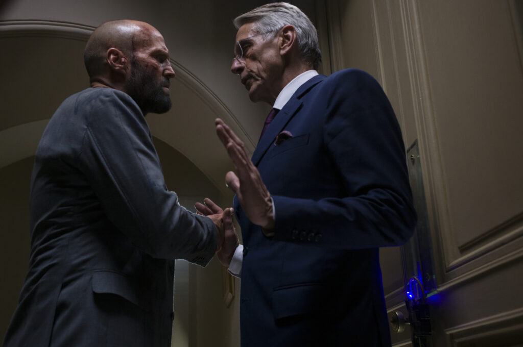 Eine spannungsgeladene Filmszene aus „The Beekeeper“ in einer Halbnahaufnahme. Auf der linken Seite steht der glatzköpfige, bärtige Hauptdarsteller Jason Statham in einem grauen Anzug im Profil und blickt grimmig auf sein Gegenüber. Rechts von ihm steht der ältere Schauspieler Jeremy Irons in einem eleganten, dunkelblauen Anzug mit Krawatte und Brille. Er hat seine linke Hand beschwichtigend mit erhobener Handfläche zwischen sich und Jason Statham ausgestreckt, während er ihn mit ernstem Blick fixiert. Die Szene spielt in einem gedimmten, eleganten Innenraum vor einer hellen Wand mit einem Rundbogen im Hintergrund.