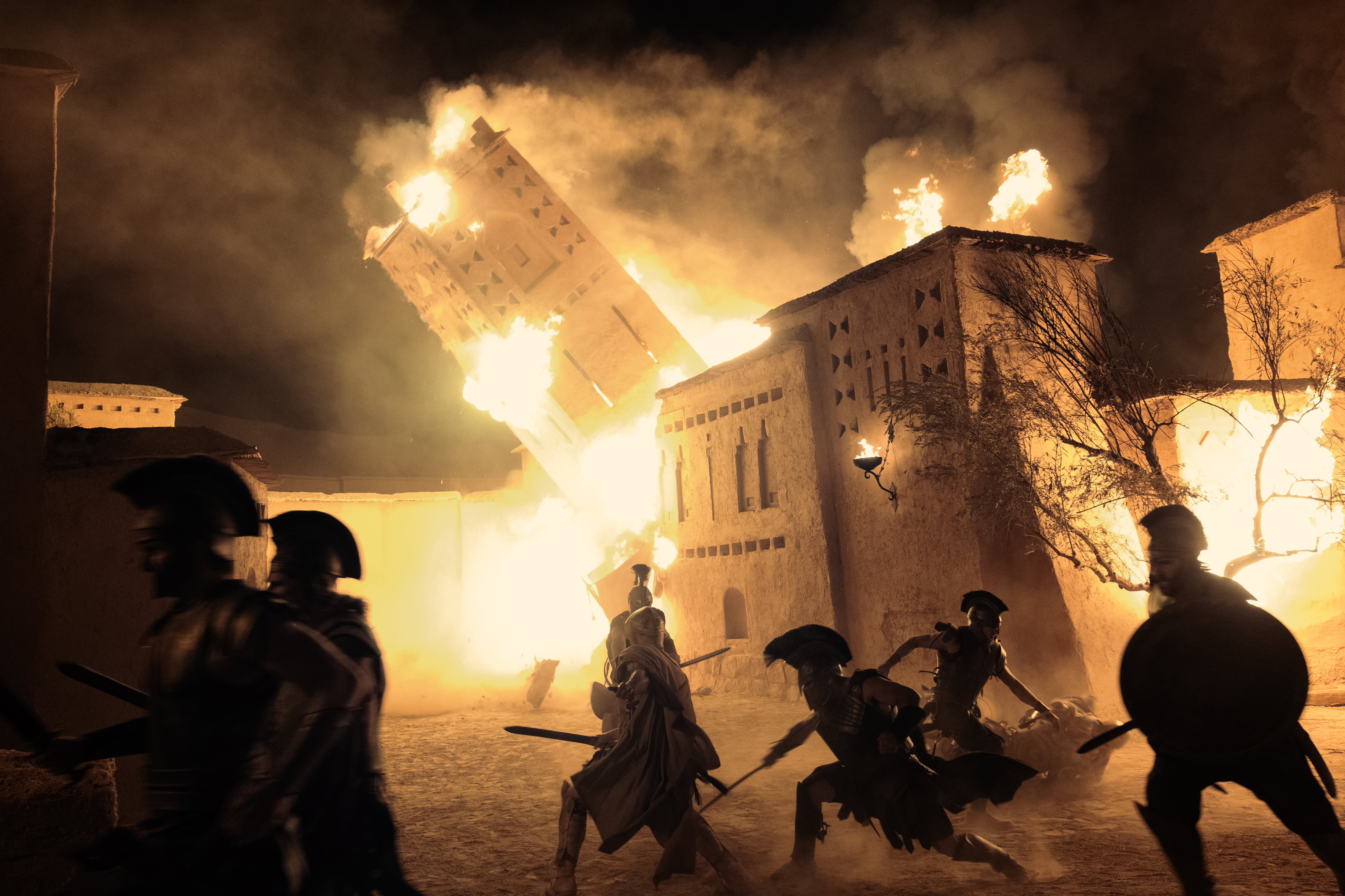 Eine dramatische, nächtliche Kriegsszene aus dem Film „Die Odyssee“. Im Hintergrund stürzt ein großer, sandfarbener antiker Festungsturm inmitten von gewaltigen Explosionen, lodernden Flammen und dichtem Rauch in Schräglage ein. Im Vordergrund sind die dunklen Silhouetten mehrerer griechischer Krieger in Rüstungen und Helmen mit Busch (Galea) zu sehen. Sie stürmen mit gezogenen Schwertern und Rundschilden im Gegenlicht des Feuers von rechts nach links durch den Innenhof der brennenden Festung.