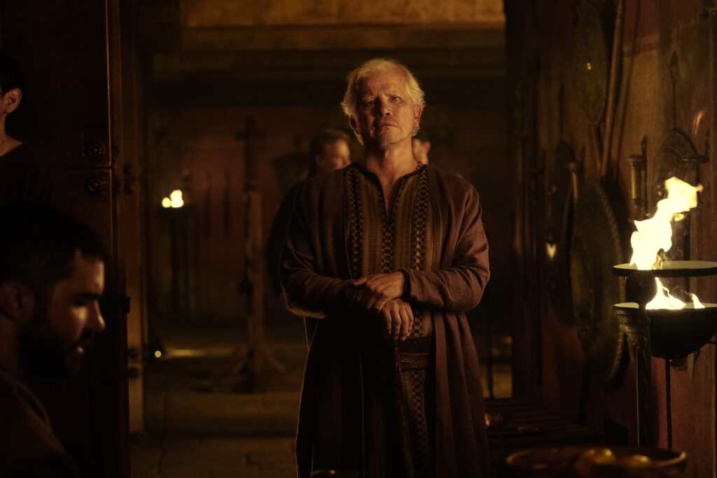 Eine düstere Filmszene aus „Die Odyssee“ mit John Leguizamo als Eumaeus. Er steht im Zentrum des Bildes in einem schwach beleuchteten, antiken Steinsaal und blickt mit ernstem, nachdenklichem Gesichtsausdruck nach vorne. Seine Hände sind vor dem Bauch zusammengelegt. Er trägt ein langes, braunes Gewand mit gemusterten Borten und hat weißes Haar sowie einen Drei-Tage-Bart. Auf der rechten Bildseite lodert eine offene Flamme in einer Metallschale (Fackelhalter), die die Szene in warmes, schattiges Licht taucht. Im linken Vordergrund ist das Profil eines anderen Mannes im Schatten zu erkennen.