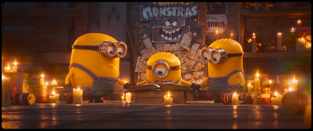 Filmszene aus Minions & Monster: Drei der kleinen gelben Kreaturen blicken im schummerigen Licht vieler brennender Kerzen auf ein magisches Buch.