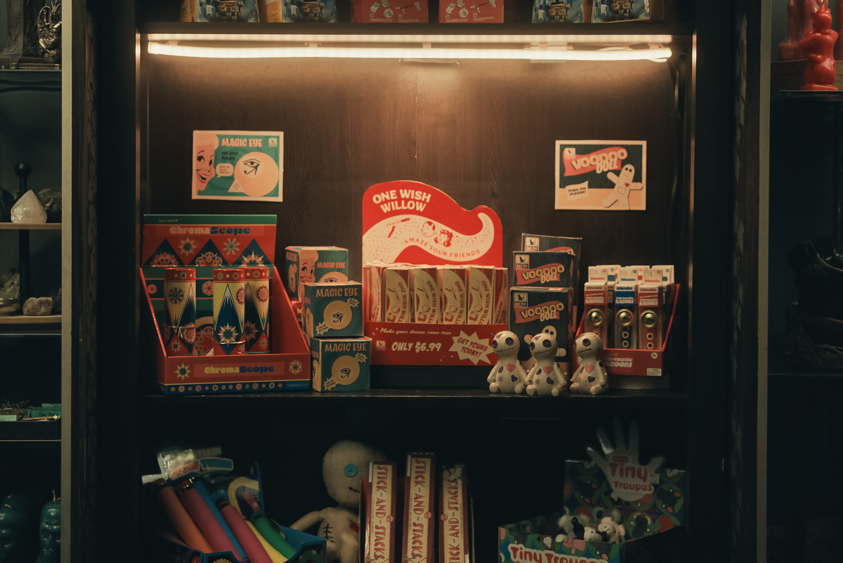 Ausschnitt aus Obsession: Der beleuchtete Ausschnitt eines Verkaufsregals mit verschiedenen Spielzeugen und Scherzartikeln im Retro-Stil. Im Zentrum steht ein rotes Display für „One Wish Willow“-Verpackungen, daneben kleine Voodoo-Puppen aus Stoff und Boxen für „Magic Eye“- sowie „ChromaScope“-Produkte. Die Atmosphäre ist durch das warme Licht der Regalleuchte und die Vintage-Verpackungen nostalgisch und leicht mystisch gestaltet.