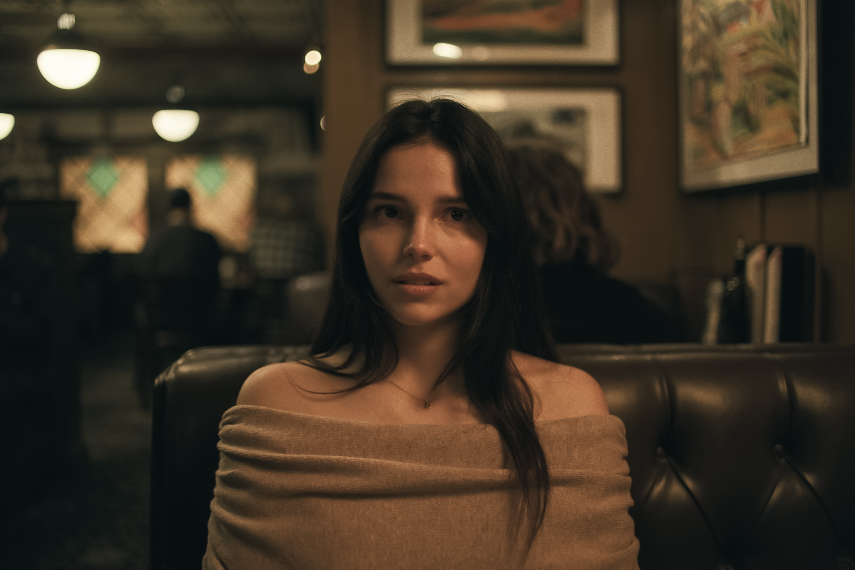 Filmszene aus dem Kinofilm „Obsession – Du sollst mich lieben“ mit der Schauspielerin Inde Navarrette als Nikki. Sie sitzt im Fokus der Kamera in einer ledernen Sitzecke eines Restaurants oder Diners. Sie trägt ein schulterfreies, hellbraunes Oberteil und blickt mit einem intensiv starrenden, leicht besorgten und emotionalen Gesichtsausdruck direkt nach vorne. Der Hintergrund ist im warmen, gedimmten Licht gehalten und zeigt unscharf angedeutete Bilderrahmen an der Wand sowie Hängelampen.