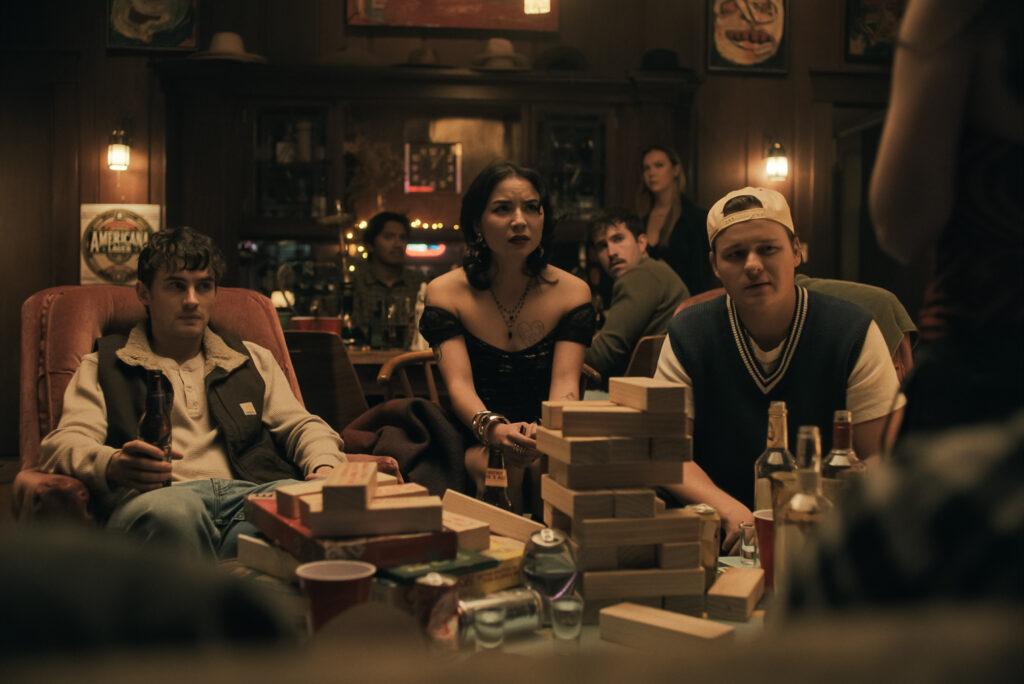 Eine Gruppe junger Erwachsener sitzt in einer schummrig beleuchteten Bar oder einem Partyraum zusammen und blickt angespannt auf ein wackeliges Holzklotz-Turmspiel (Jenga) auf dem Tisch im Vordergrund. Drei Personen sitzen im Fokus auf Sesseln und Stühlen, umgeben von Getränkeflaschen, Dosen und Bechern, während im Hintergrund weitere Personen und die rustikale Holzbar zu sehen sind. Die Stimmung wirkt konzentriert und erwartungsvoll.
