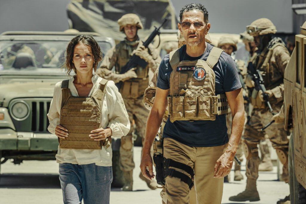 Filmszene aus dem Kriegsdrama „Operation: Kabul“ in einer Halbnahaufnahme. Im Vordergrund laufen eine junge Frau mit erschöpftem, besorgtem Gesichtsausdruck und ein Mann mit Sonnenbrille und ernster Miene nebeneinander her. Die Frau trägt eine helle Bluse und eine braune ballistische Schutzweste. Der Mann, gespielt von Roschdy Zem, trägt ein dunkles T-Shirt unter einer militärischen Weste mit runden Abzeichen. Im staubigen Hintergrund sind mehrere bewaffnete Soldaten in Wüstentarn-Ausrüstung vor einem Militärfahrzeug zu sehen.