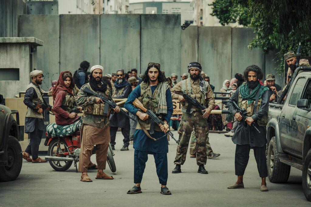 Filmszene aus dem Kriegsdrama „Operation: Kabul“. Eine schwer bewaffnete Gruppe von Taliban-Kämpfern steht aufgereiht inmitten einer staubigen Straße in Kabul, umgeben von hohen, grauen Schutzwänden aus Beton. Im Zentrum steht ein junger Kämpfer mit langen, dunklen Haaren, einer traditionellen Kopfbedeckung (Pakol) und einer Militärweste, der eine Kalaschnikow (AK-47) schussbereit mit beiden Händen vor dem Körper hält. Flankiert wird er von weiteren bärtigen Männern in traditionellen Gewändern und Tarnkleidung, die ebenfalls Sturmgewehre tragen. Auf der rechten Bildseite parkt ein staubiger Pickup-Truck, während im Hintergrund eine unruhige Menschenmenge hinter einer Absperrung zu erkennen ist.