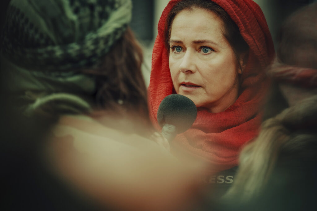 Eine dramatische Nahaufnahme aus dem Film „Operation: Kabul“ mit der Schauspielerin Sidse Babett Knudsen. Sie steht im rechten Bildhintergrund, trägt ein leuchtend rotes Kopftuch und blickt mit einem besorgten, emotionalen Gesichtsausdruck und geweiteten blauen Augen nach links. Direkt vor ihr ist ein dunkles Handmikrofon mit Schaumstoffschutz zu sehen. Auf ihrer Kleidung ist der Schriftzug „PRESS“ zu erahnen. Der linke Vordergrund zeigt die unscharf verschwommene Silhouette einer anderen Person mit einem grünen Kopftuch, was der Szene eine dichte, bedrückende Atmosphäre verleiht.