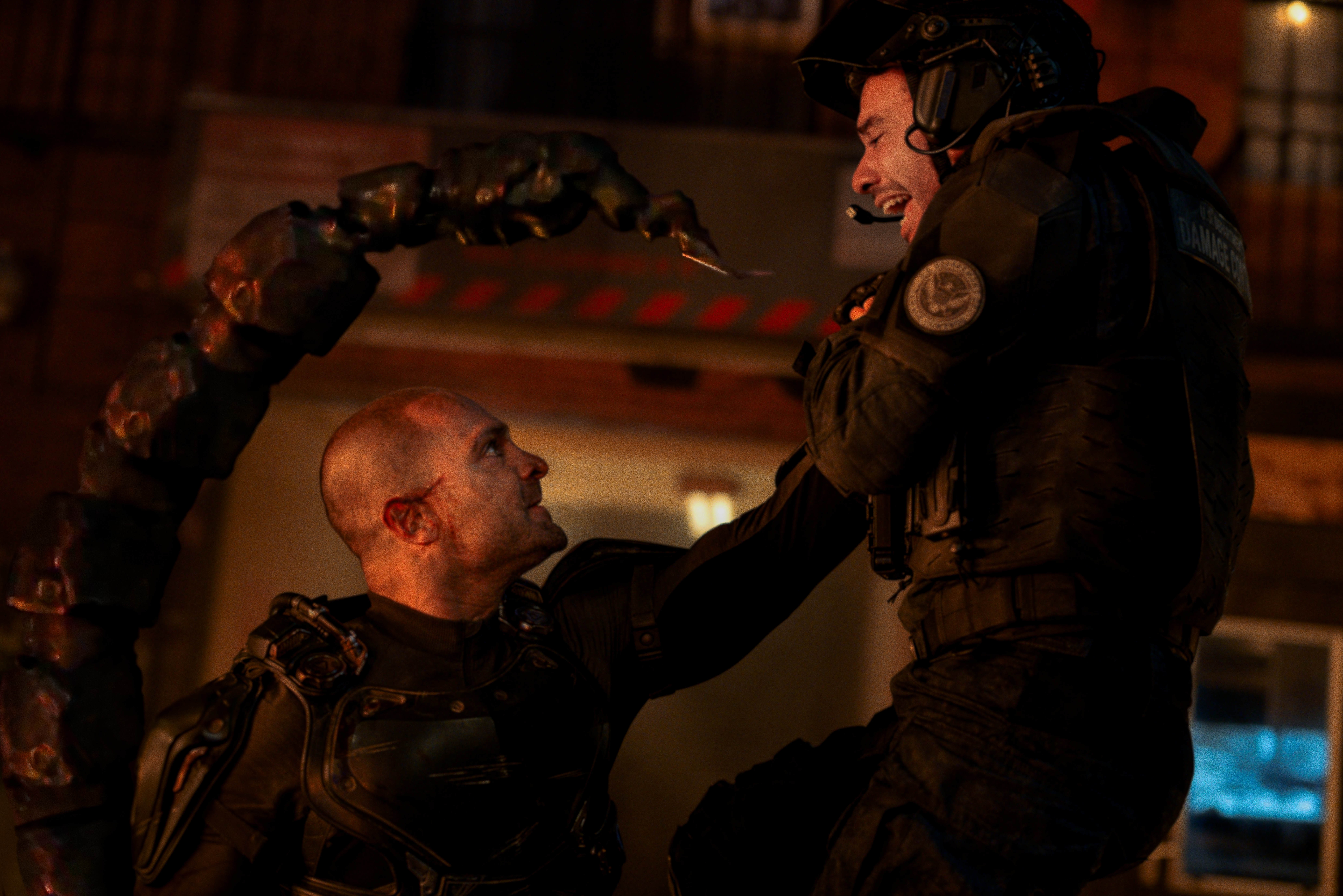 Filmszene aus Spider-Man: Brand New Day, in der der Schurke Scorpion (Michael Mando) in einem schwarzen Tech-Anzug einen schwer bewaffneten Soldaten der Regierungsbehörde Damage Control mit einer Hand in die Luft hebt. Im Vordergrund windet sich Scorpions mechanischer Stachelschwanz nach oben, während im Hintergrund eine nächtliche, rötlich beleuchtete Kulisse zu sehen ist.