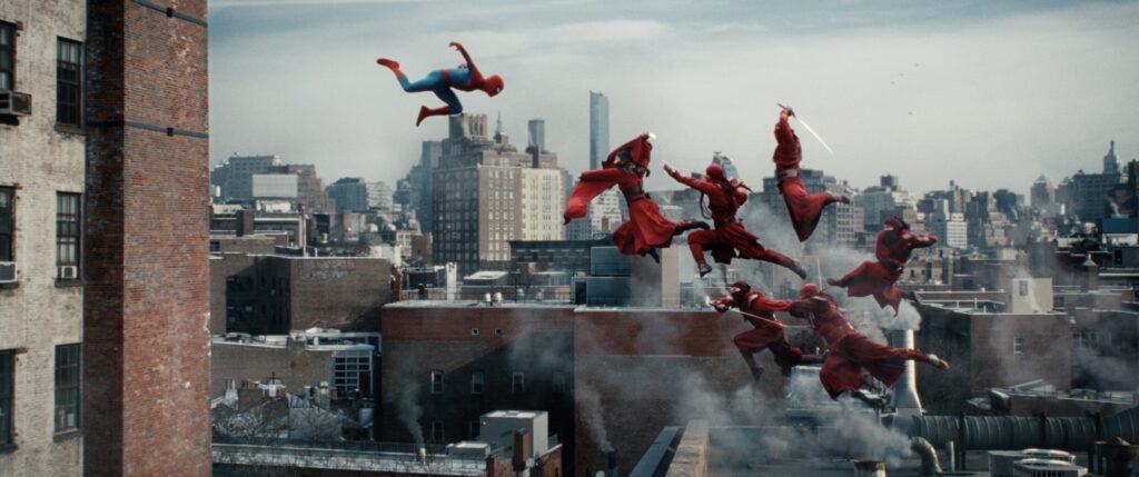 Weitwinkel-Filmszene über den Dächern von New York, in der Spider-Man in der Luft über einer Gruppe von sechs rot gekleideten Ninja-Kämpfern der „Hand“ schwebt. Die Ninjas springen mit gezogenen Schwertern dynamisch von einem Hausdach ab, während im Hintergrund die dichte Skyline von Manhattan unter einem bewölkten Himmel zu sehen ist.