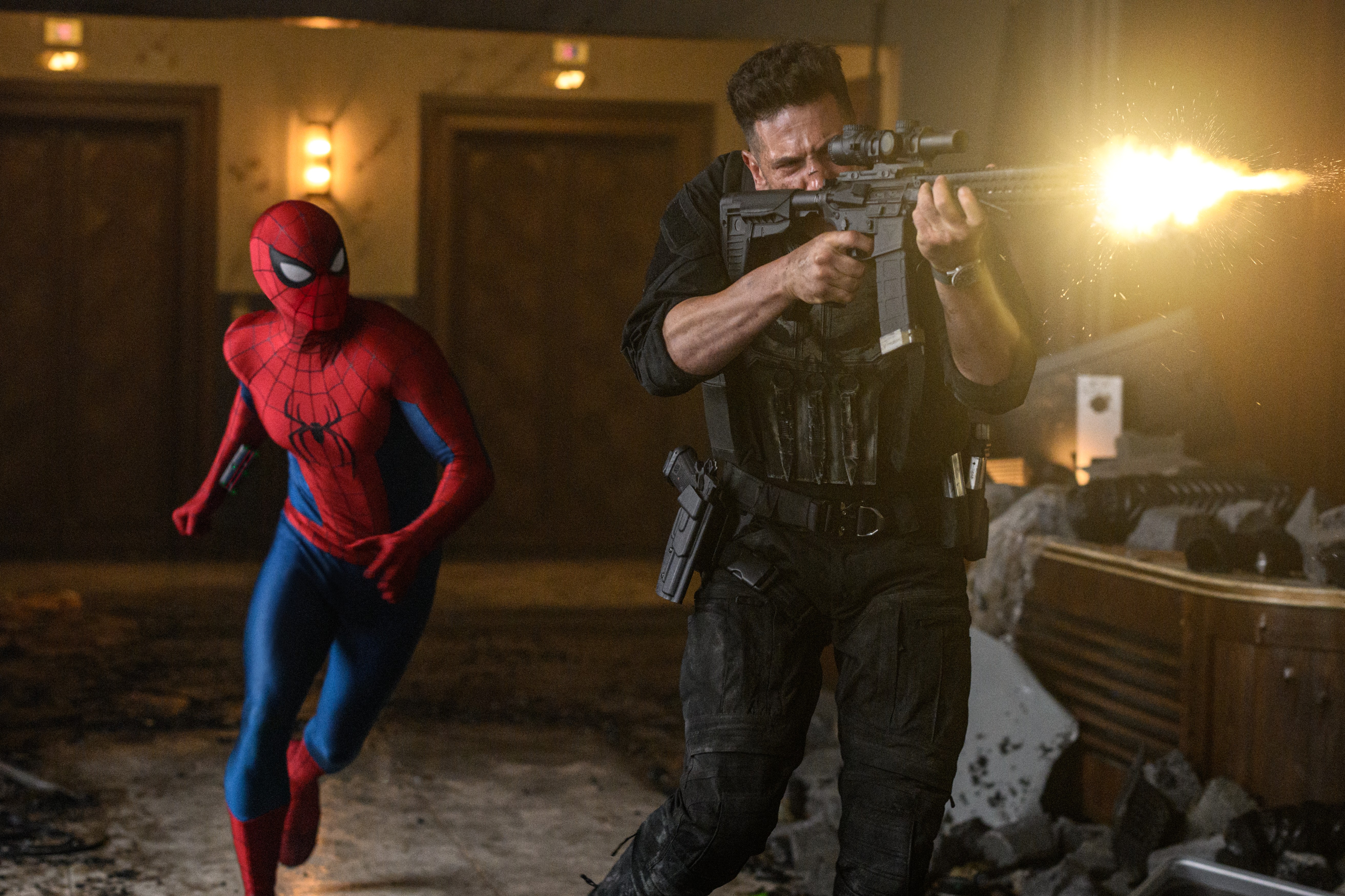 Actionreiche Filmszene in einem zerstörten Innenraum, in der The Punisher (Jon Bernthal) im Vordergrund in taktischer, schwarzer Kleidung ein Sturmgewehr abfeuert, aus dessen Lauf ein helles Mündungsfeuer bricht. Links hinter ihm läuft Spider-Man im klassisch rot-blauen Kostüm in geduckter Haltung durch den rauchigen Raum.