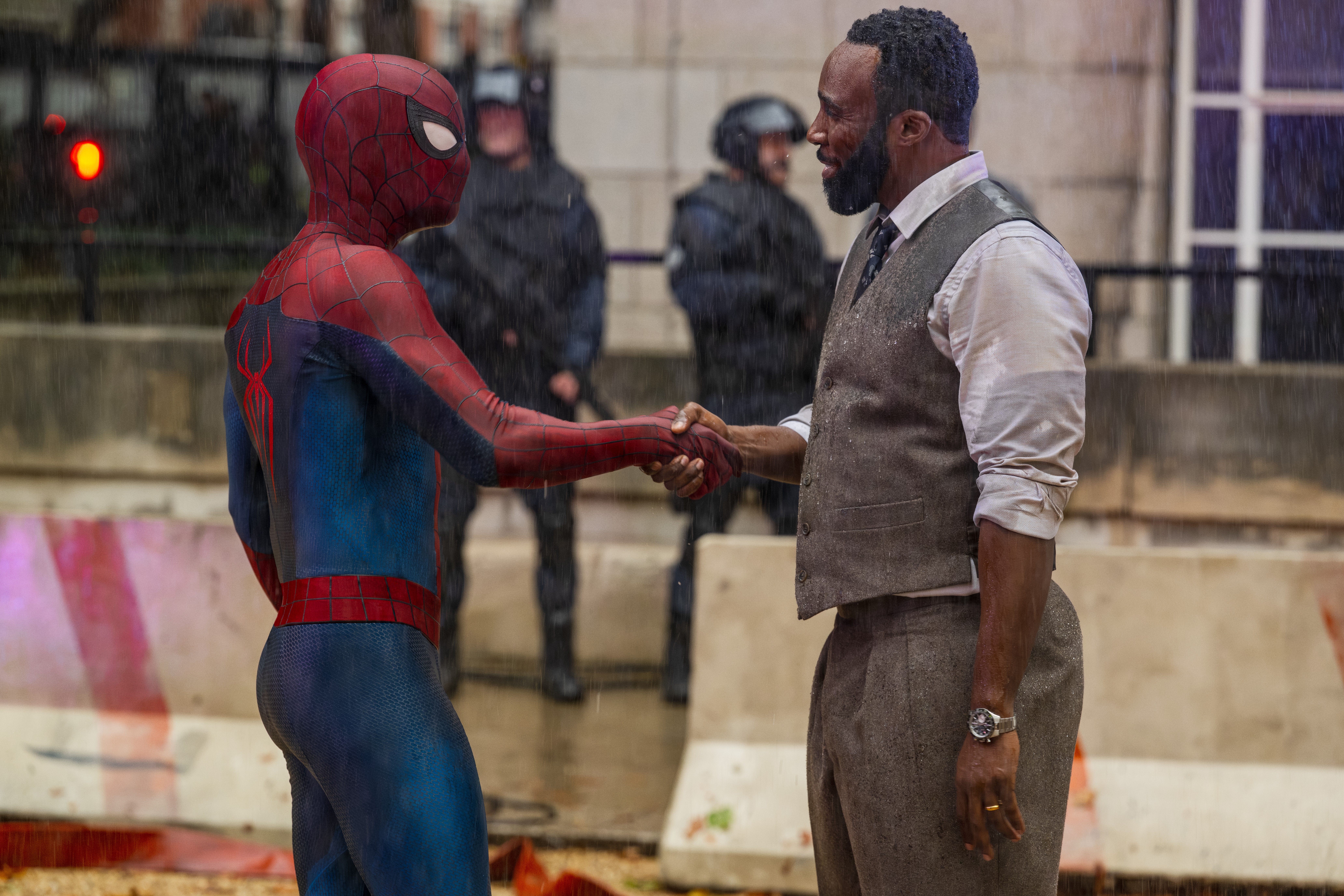 Filmszene im strömenden Regen, in der Spider-Man in seinem klassisch rot-blauen Kostüm von links kommend William Metzger (gespielt von Tramell Tillman) im Profil die Hand schüttelt. Der bärtige Mann trägt ein nasses weißes Hemd, eine graue Weste und eine passende Hose. Im unscharfen Hintergrund stehen zwei schwer bewaffnete Polizisten in taktischer Ausrüstung hinter einer Absperrung.