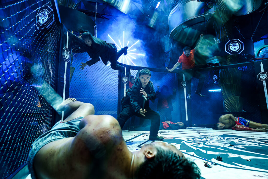 Szenenbild aus „The Furious“: Mehrere Männer in einer dynamischen, chaotischen Kampfsportszene inmitten eines von Maschendraht umzäunten MMA-Käfigs bei kühlem, blauem Scheinwerferlicht.