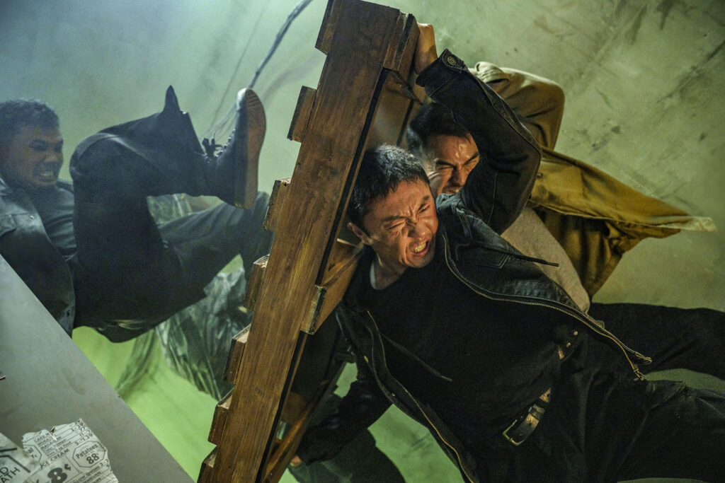Szenenbild aus „The Furious“: Hauptdarsteller Xie Miao und Joe Taslim drücken mit voller Anstrengung eine schwere Holzpalette gegen einen Angreifer in einem düsteren Raum.