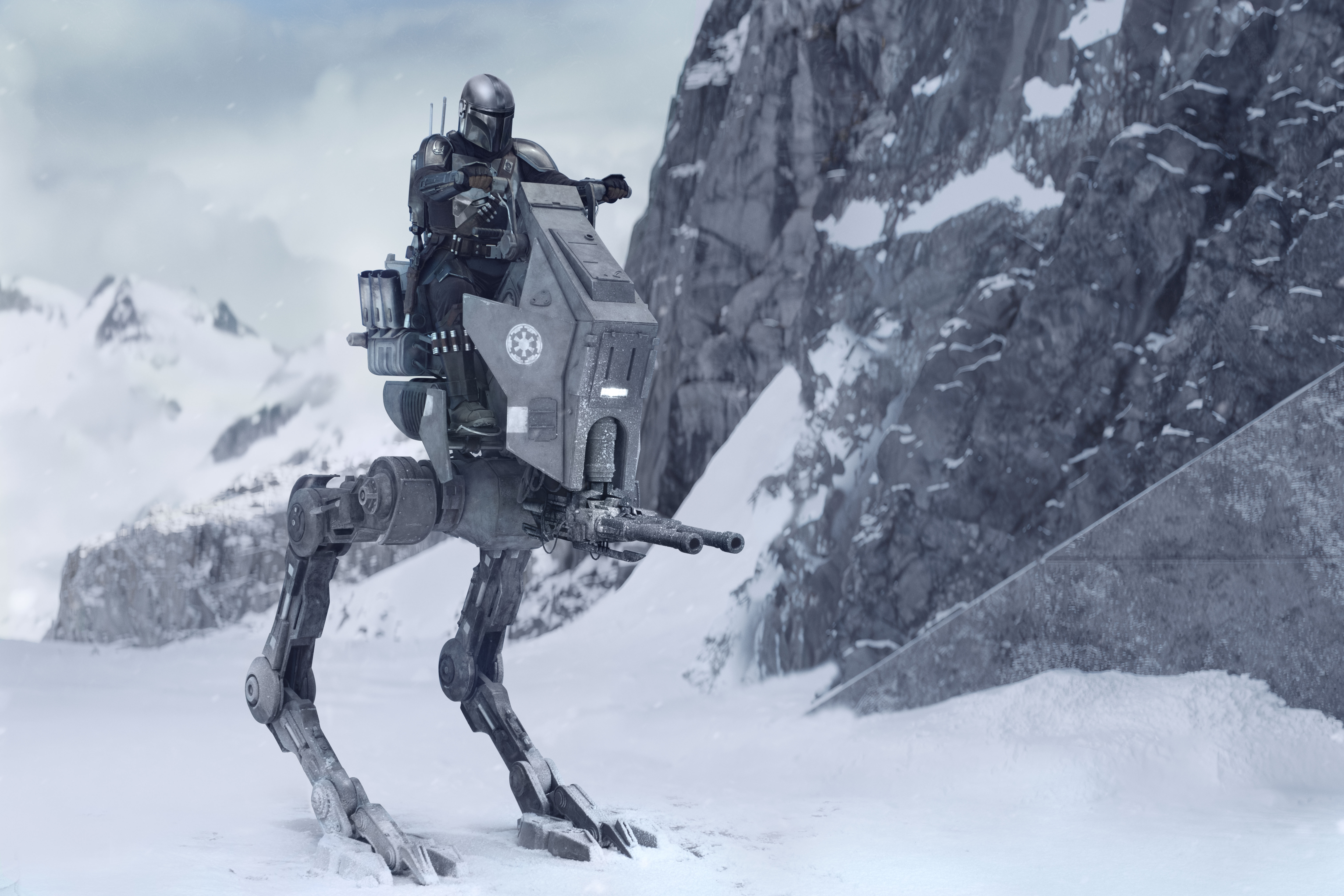 Szenenbild aus Star Wars: The Mandalorian and Grogu: Eine Person in der typischen metallischen Mandalorianer-Rüstung inklusive Helm sitzt auf einem grauen, mechanischen AT-RT-Kampfläufer. Das Fahrzeug steht auf einer schneebedeckten Ebene vor einer massiven, grau-weißen Felswand im Hintergrund. Das Wetter wirkt bewölkt und winterlich.