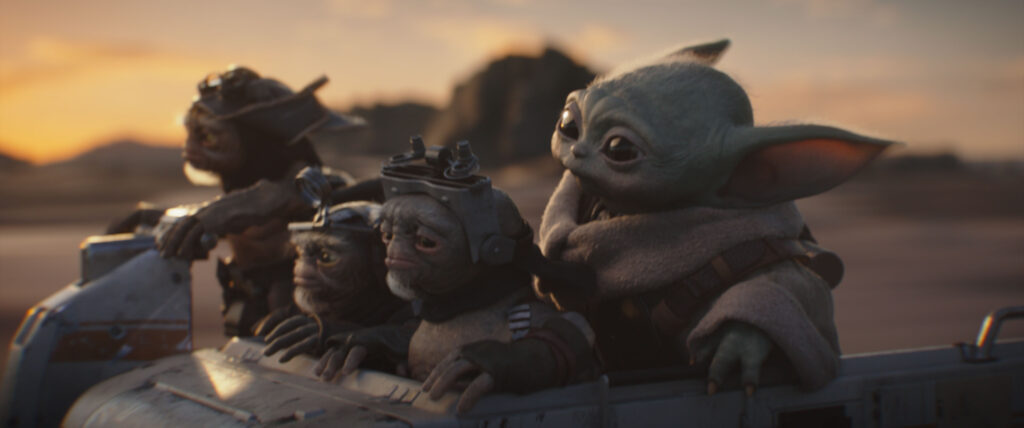 Nahaufnahme einer Filmszene aus Star Wars: The Mandalorian and Grogu: Das grüne, großohrige Wesen Grogu blickt aufmerksam nach vorne, während er in einem metallischen Fahrzeug sitzt. Neben ihm befinden sich drei sehr kleine, behaarte Kreaturen mit Schnurrbärten und Schweißerbrillen (Anzellaner). Im Hintergrund erstreckt sich eine unscharfe Wüstenlandschaft im warmen Licht des Sonnenuntergangs.