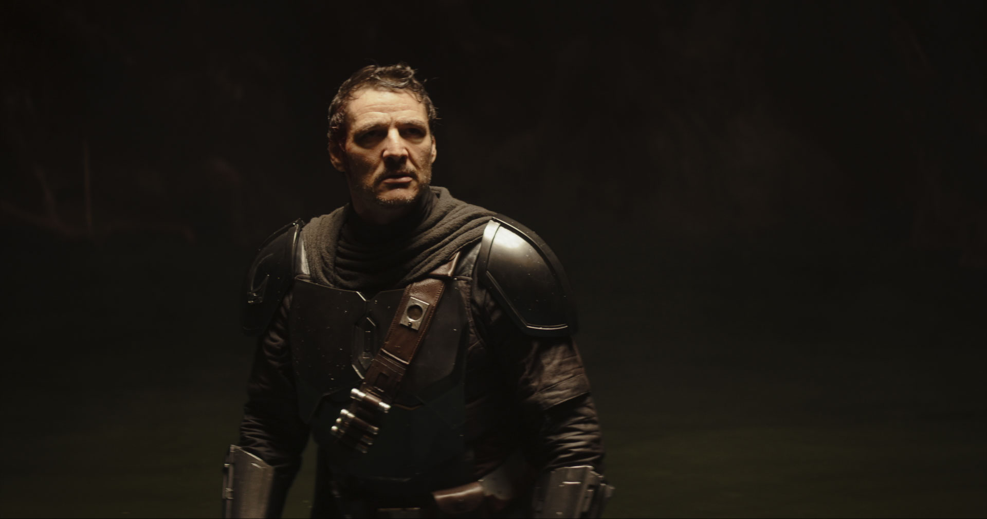 Porträtaufnahme von Pedro Pascal in der Rolle des Mandalorianers Din Djarin. Er trägt seine dunkle Beskar-Rüstung mit Schulterplatten und Munitionsgurt, hat jedoch den Helm abgenommen. Sein Blick ist ernst und leicht zur Seite gerichtet. Die Szene ist spärlich von der Seite beleuchtet und der Hintergrund versinkt in tiefer Dunkelheit.