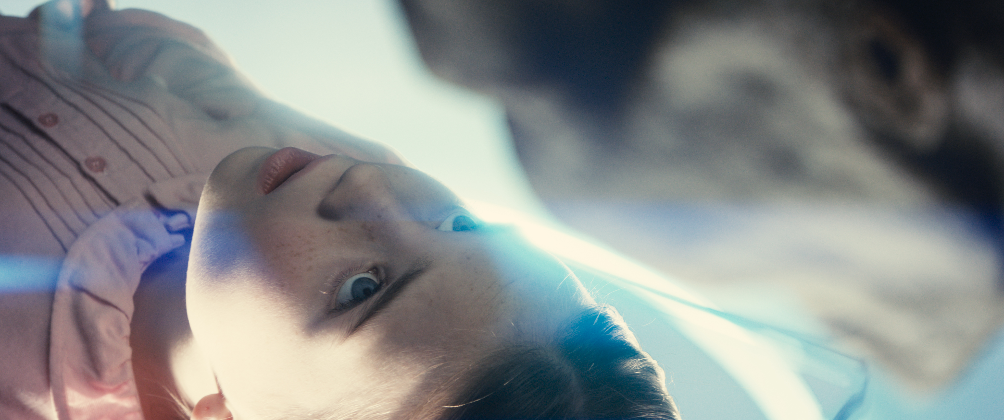 Szenenbild aus Steven Spielbergs Science-Fiction-Thriller „Disclosure Day – Der Tag der Wahrheit“ (2026). Die Kamera blickt aus einer extremen Untersicht von unten durch eine Glasscheibe nach oben. Zu sehen ist das Gesicht eines jungen Mädchens mit Sommersprossen und einer hellrosa Bluse, das mit großen, besorgten Augen direkt nach unten blickt. Ein bläulicher Lichtstrahl zieht sich quer über ihr Gesicht.