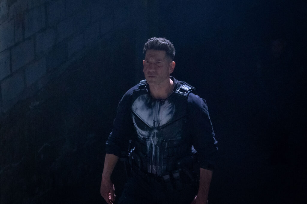 Eine düstere Aufnahme des Schauspielers Jon Bernthal als Frank Castle (The Punisher). Er trägt seine schwarze Einsatztaktik-Weste mit dem markanten, weißen Totenkopf-Logo auf der Brust. Er blickt mit ernstem, entschlossenem Gesichtsausdruck leicht zur Seite. Der Hintergrund ist eine dunkle, steinerne Wand im Halbschatten.