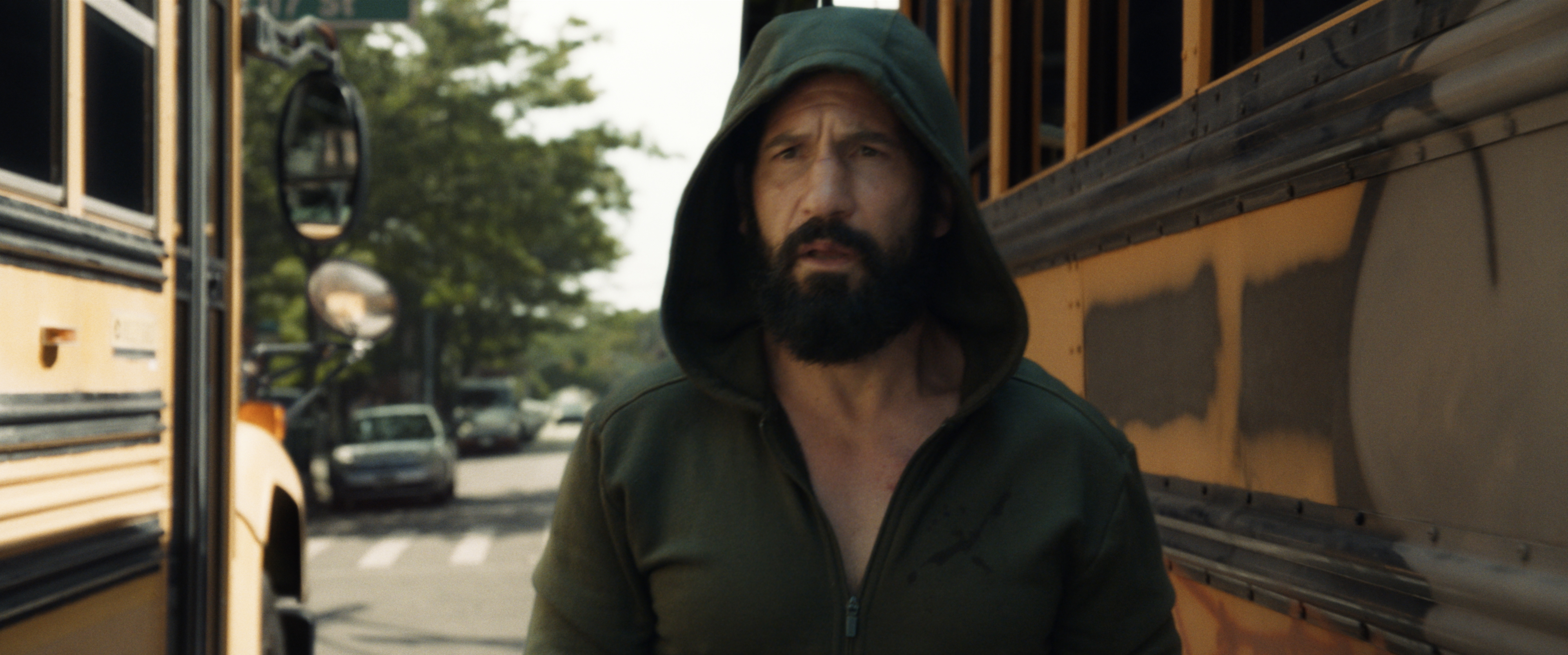 Ein Bild aus The Punisher: One Last Kill: Nahaufnahme des Schauspielers Jon Bernthal als Frank Castle (The Punisher) mit dichtem, schwarzem Vollbart. Er blickt mit ernstem, erschöpftem Gesichtsausdruck direkt nach vorne. Er trägt eine tief ins Gesicht gezogene, dunkelgrüne Kapuzenjacke. Im Hintergrund steht er auf einer Straße in New York City zwischen zwei gelben Schulbussen.